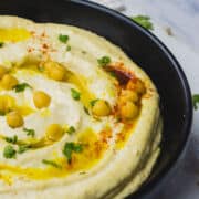 A black bowl filled with creamy hummus dip, topped with whole chickpeas, olive oil, chopped parsley, and a sprinkle of paprika, sits on a white surface with a striped towel nearby.