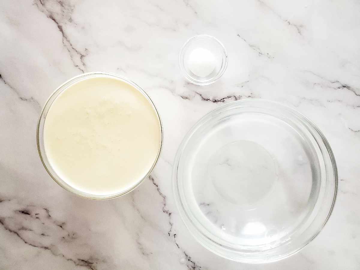 Three clear bowls on a white marble surface: one medium bowl with heavy cream, one large bowl of ice water and a small bowl of plain yogurt.