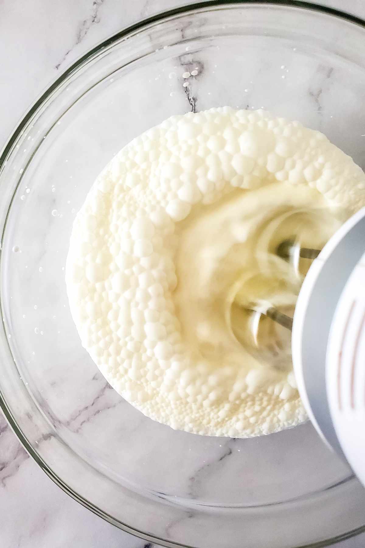 A hand mixer is whipping cream in a glass bowl, creating bubbles and froth on the surface—a process that can lead to rich cultured butter. The background is a white marble countertop.
