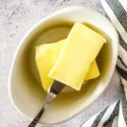 A small white dish with two rectangular slices of cultured butter, one of which is held by a butter knife. A striped cloth is partially visible to the right on a light textured surface.
