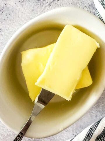 A small white dish with two rectangular slices of cultured butter, one of which is held by a butter knife. A striped cloth is partially visible to the right on a light textured surface.