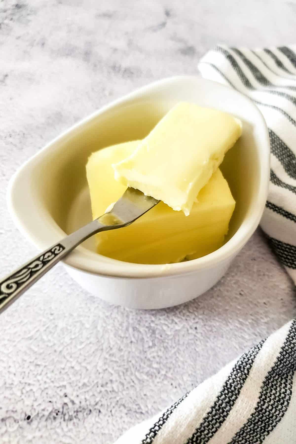 A ceramic dish holds two pieces of cultured butter, one speared with a decorative fork.