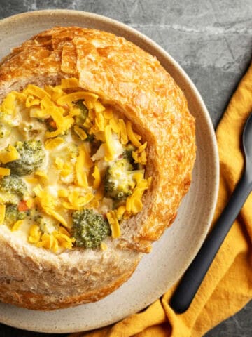 A bread bowl filled with rich broccoli cheddar soup, topped with shredded cheddar, sits on a plate next to a black spoon and a yellow cloth napkin on a gray surface.