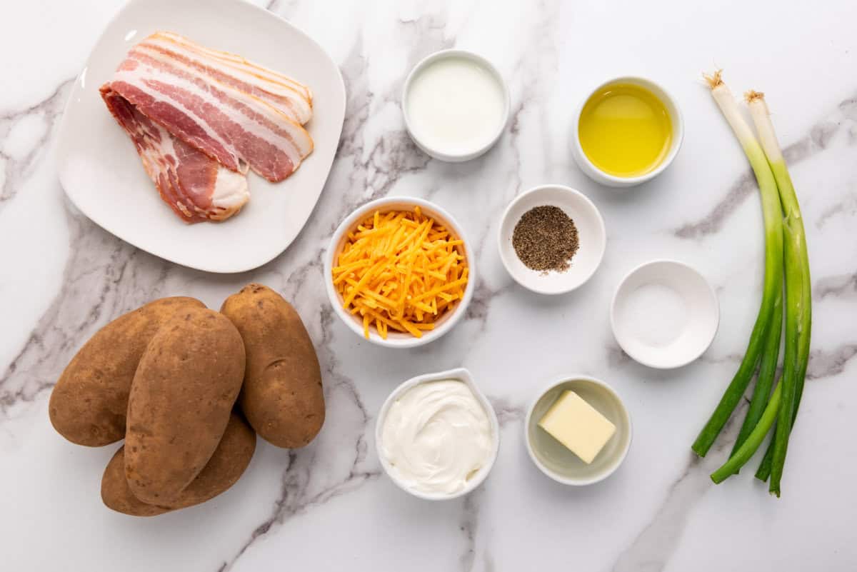 A marble surface with ingredients laid out for easy twice baked potatoes: three russet potatoes, strips of bacon, shredded cheddar cheese, sour cream, butter, milk, olive oil, black pepper, salt in small bowls, and two green onions.