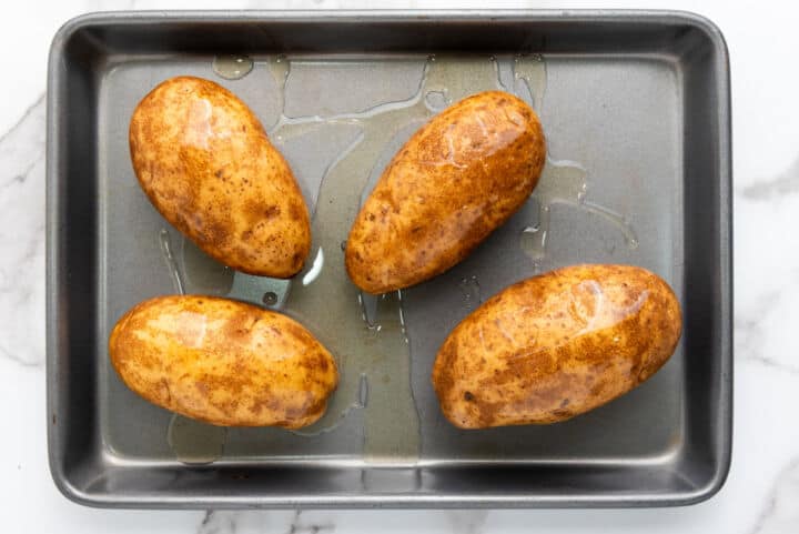 Four whole russet potatoes with brown skins, coated in oil, rest on a metal baking sheet, ready for the oven. Perfect for making easy twice baked potatoes, they sit atop a white marble countertop.