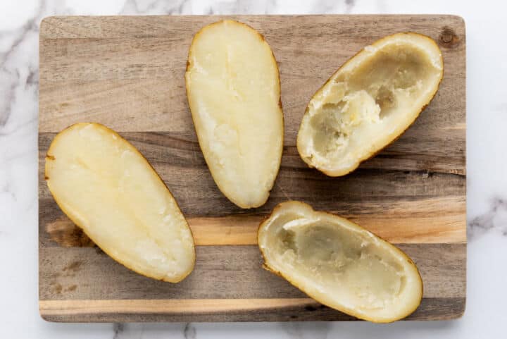 Four potato halves on a wooden cutting board, perfect for making easy twice baked potatoes—two with most of the insides scooped out and two mostly intact, all placed on a marble surface.