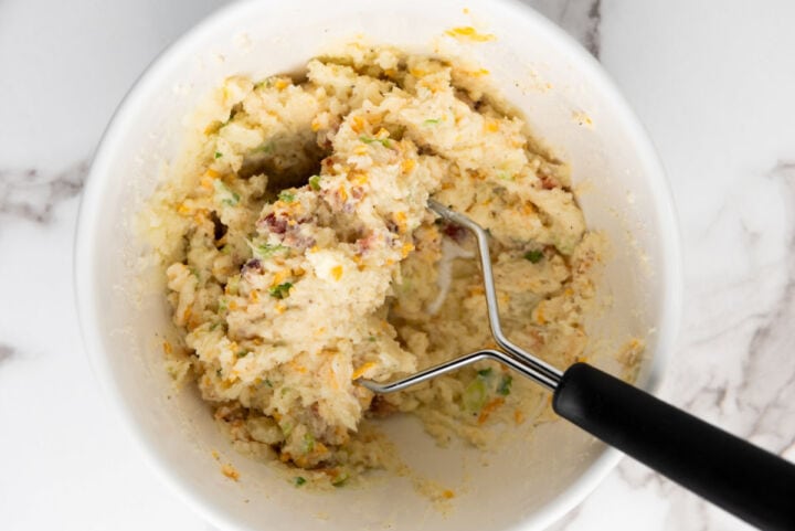 A bowl of mashed potatoes mixed with bacon, cheese, and green onions-just like the filling for easy twice baked potatoes-being mashed with a black-handled potato masher on a white marble surface.