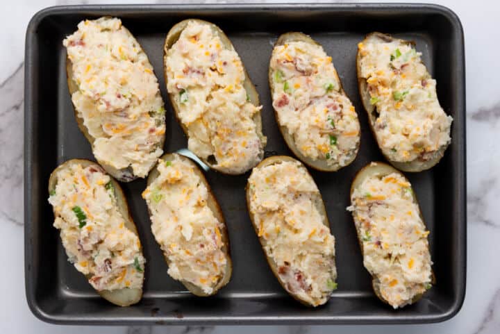 A baking tray with eight halved potatoes filled with a creamy mixture of cheese, bacon bits, and green onions—perfect for making easy twice baked potatoes.