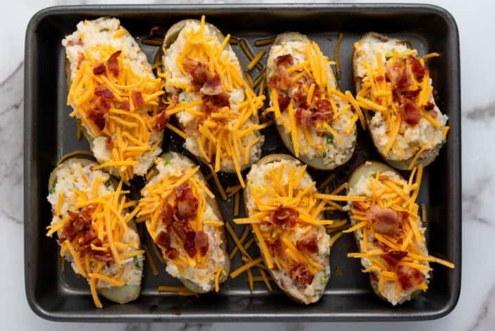 A baking tray filled with easy twice baked potatoes-halves topped with shredded cheddar cheese, crumbled bacon, and green onions-ready to be baked.