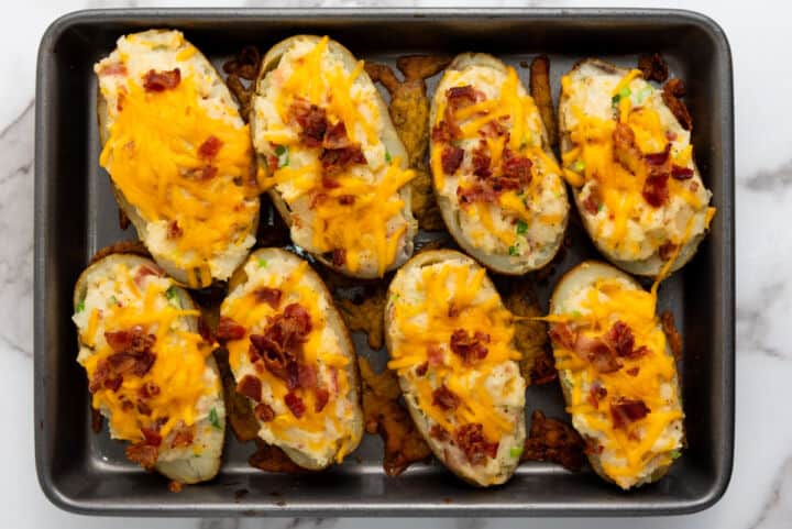 A baking tray filled with eight easy twice baked potatoes, each topped with melted cheddar cheese, bacon bits, and chopped green onions on a white marble surface.