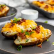 Easy twice baked potatoes-two crispy halves topped with melted cheddar, bacon bits, and green onions-are served on a gray plate, with a small dish of sour cream in the background.