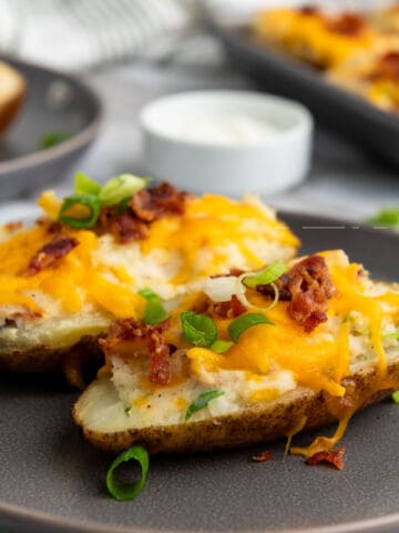 Easy twice baked potatoes-two crispy halves topped with melted cheddar, bacon bits, and green onions-are served on a gray plate, with a small dish of sour cream in the background.
