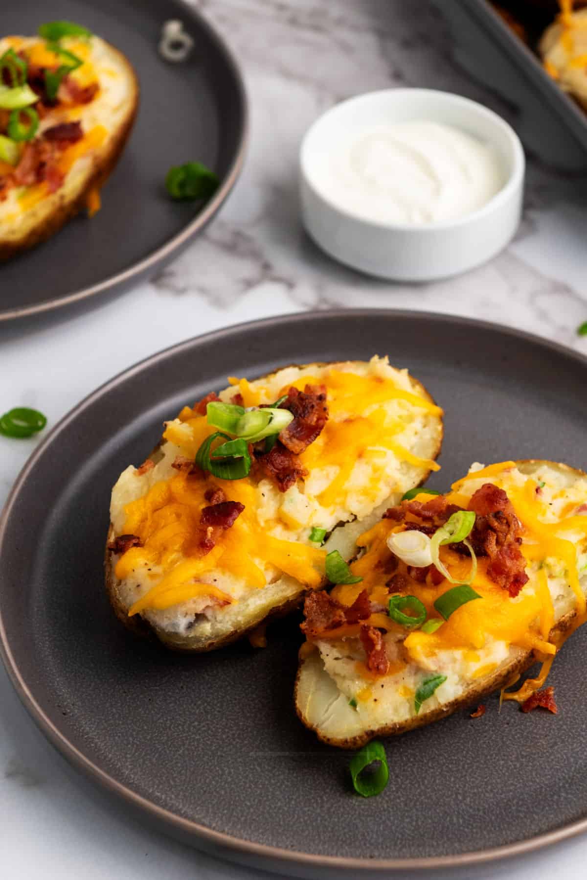 Two easy twice baked potatoes on a dark plate, topped with melted cheddar cheese, crispy bacon bits, and sliced green onions. A small bowl of sour cream sits in the background on a marble surface.