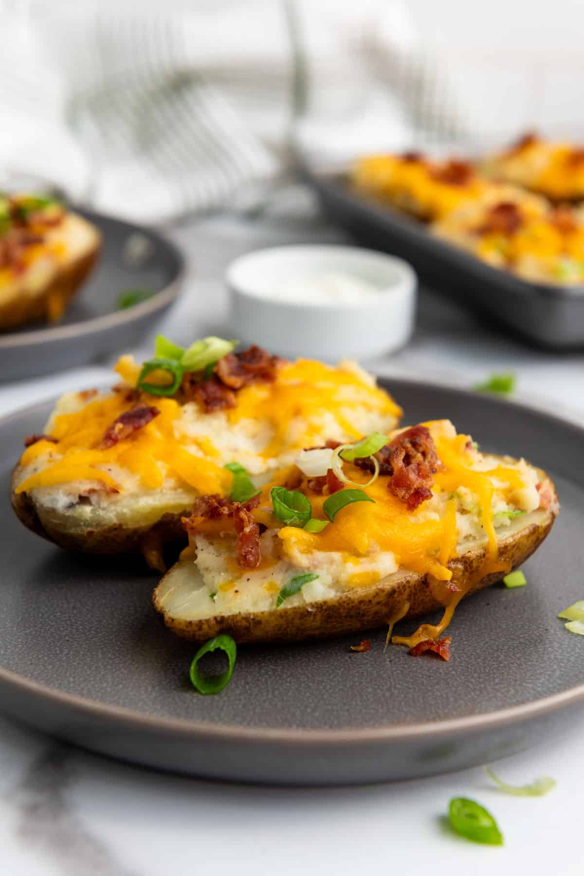 Easy twice baked potatoes: Two potato halves topped with melted cheddar cheese, bacon bits, and green onions on a gray plate, with more potatoes and a small bowl of sour cream in the background.