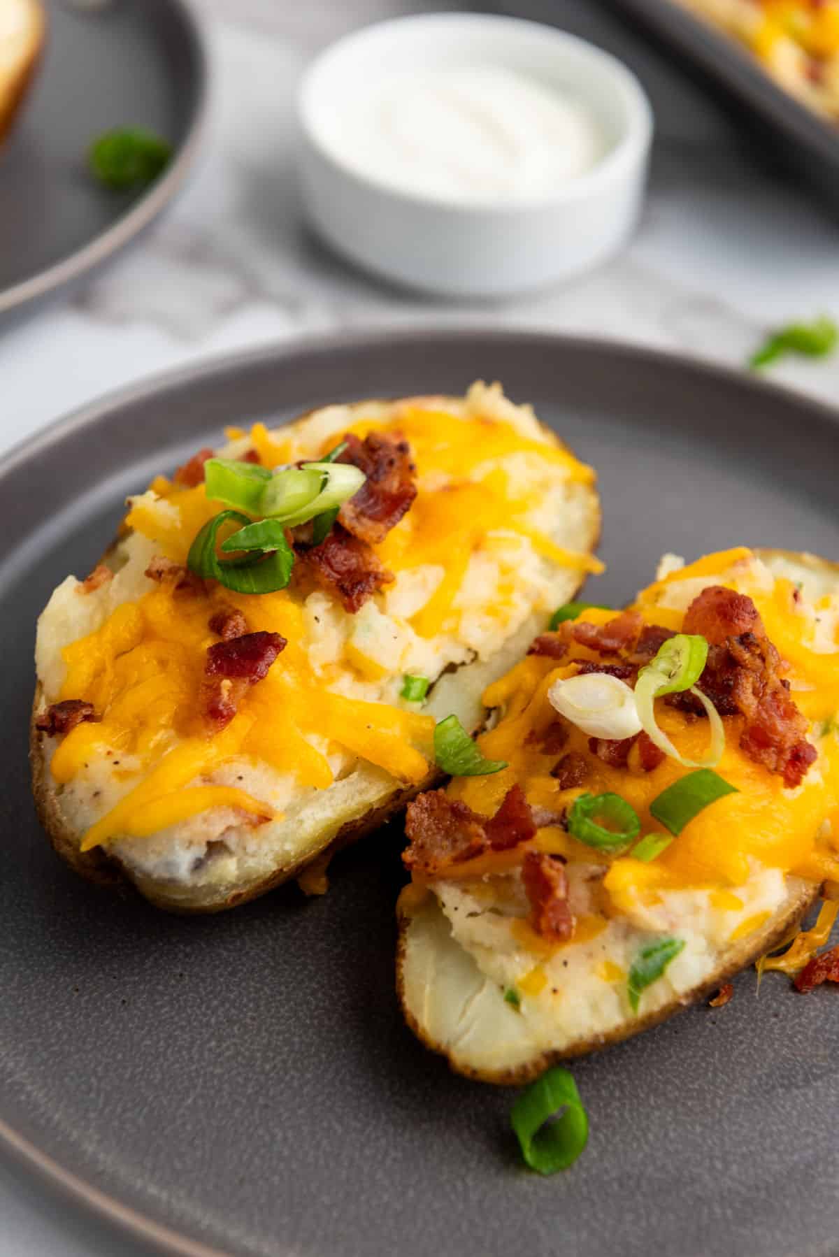 Two easy twice baked potatoes sit on a gray plate, topped with melted cheddar cheese, crispy bacon pieces, and sliced green onions. A small bowl of sour cream is in the background.