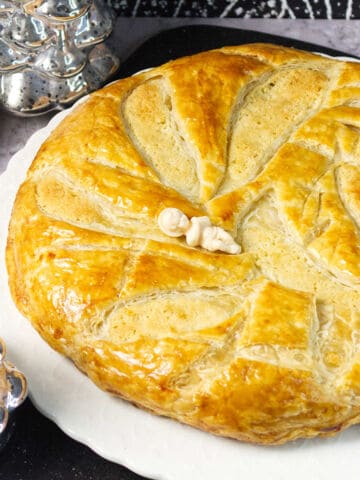 A golden, round puff pastry cake inspired by the classic galette des rois recipe sits on a white plate, decorated with festive slits and patterns. A small white figurine tops the pastry, with silver holiday tree ornaments nearby.