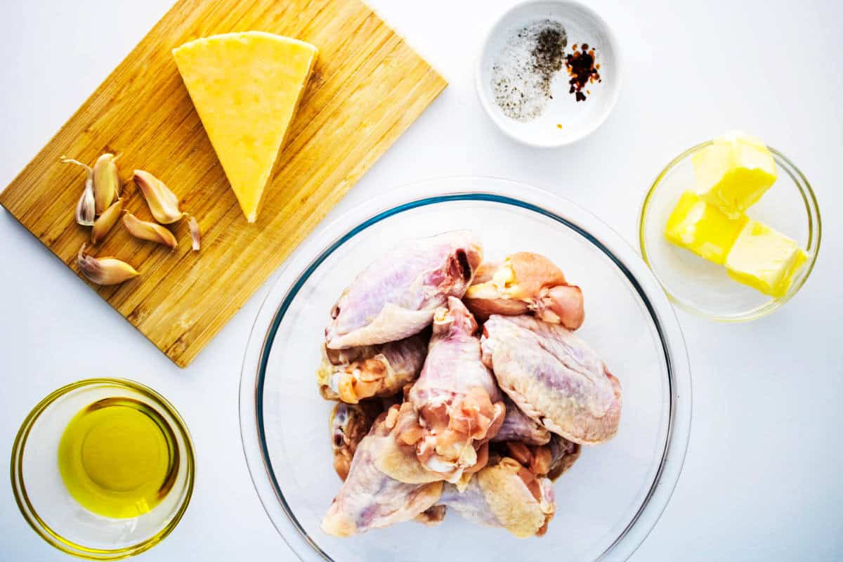 A glass bowl of raw wings, perfect for garlic parmesan chicken wings, is surrounded by olive oil, garlic cloves, cheddar cheese on a wooden board, plus a small bowl of spices and butter cubes, all set on a white surface.