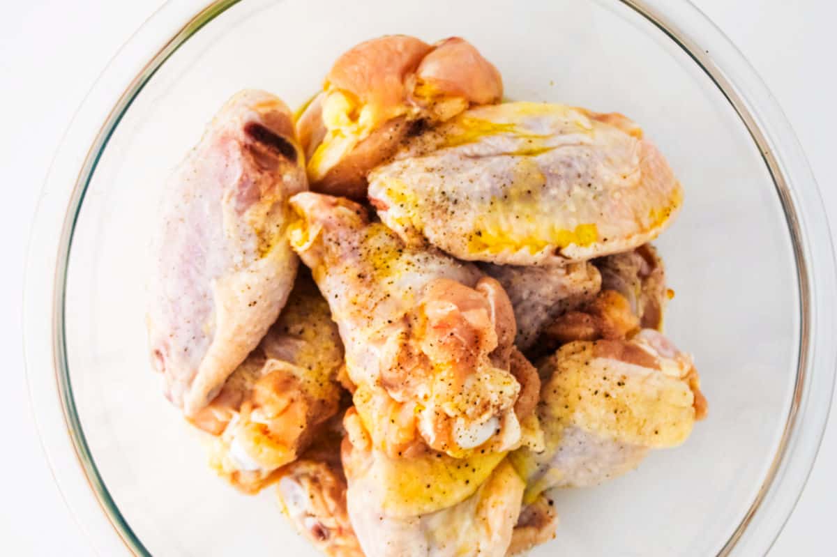A glass bowl filled with raw chicken wings, lightly seasoned with salt, pepper, and hints of garlic parmesan, on a white background.