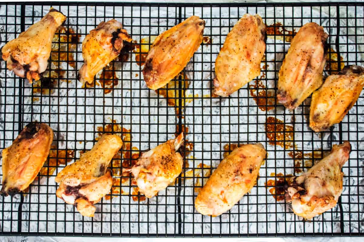 Twelve seasoned, baked garlic parmesan chicken wings are arranged on a black wire rack set over a foil-lined baking sheet. The wings are golden brown, with small spots of dripped oil underneath.