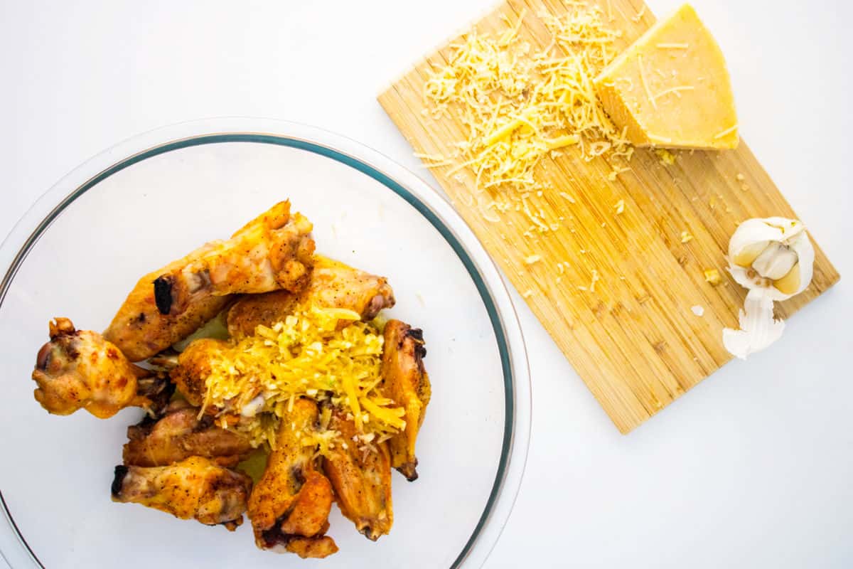 A plate of garlic parmesan chicken wings topped with shredded cheese sits next to a wooden cutting board with a block of cheese, cheese shreds, and a garlic bulb.