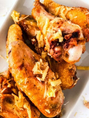 A close-up of crispy garlic parmesan chicken wings on a white plate, topped with shredded cheese and seasonings.