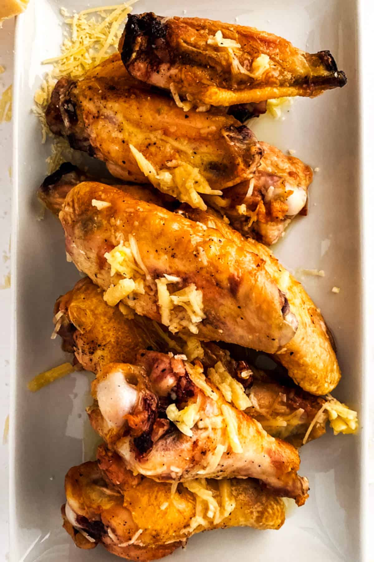 A close-up of several garlic parmesan chicken wings arranged on a white rectangular plate, topped with shredded cheese.