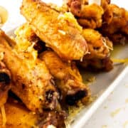 A close-up of garlic parmesan chicken wings stacked on a white plate, garnished with grated cheese. The wings are golden brown and juicy, with a piece of cheese visible in the foreground.