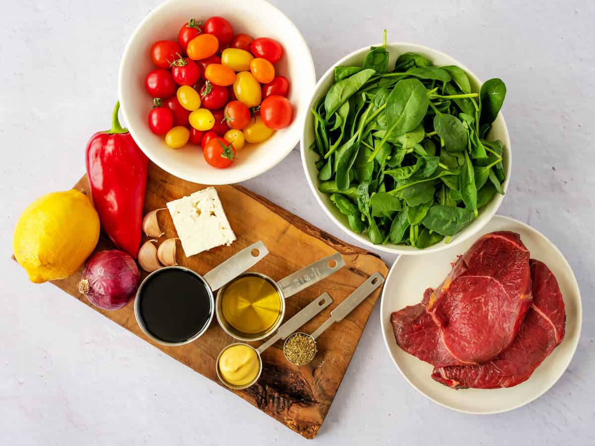 A selection of fresh ingredients on a table for a grilled steak salad: bowls of cherry tomatoes, spinach, raw beef steaks, a lemon, red pepper, red onion, garlic, feta cheese, mustard, dried herbs, olive oil, and balsamic vinegar.