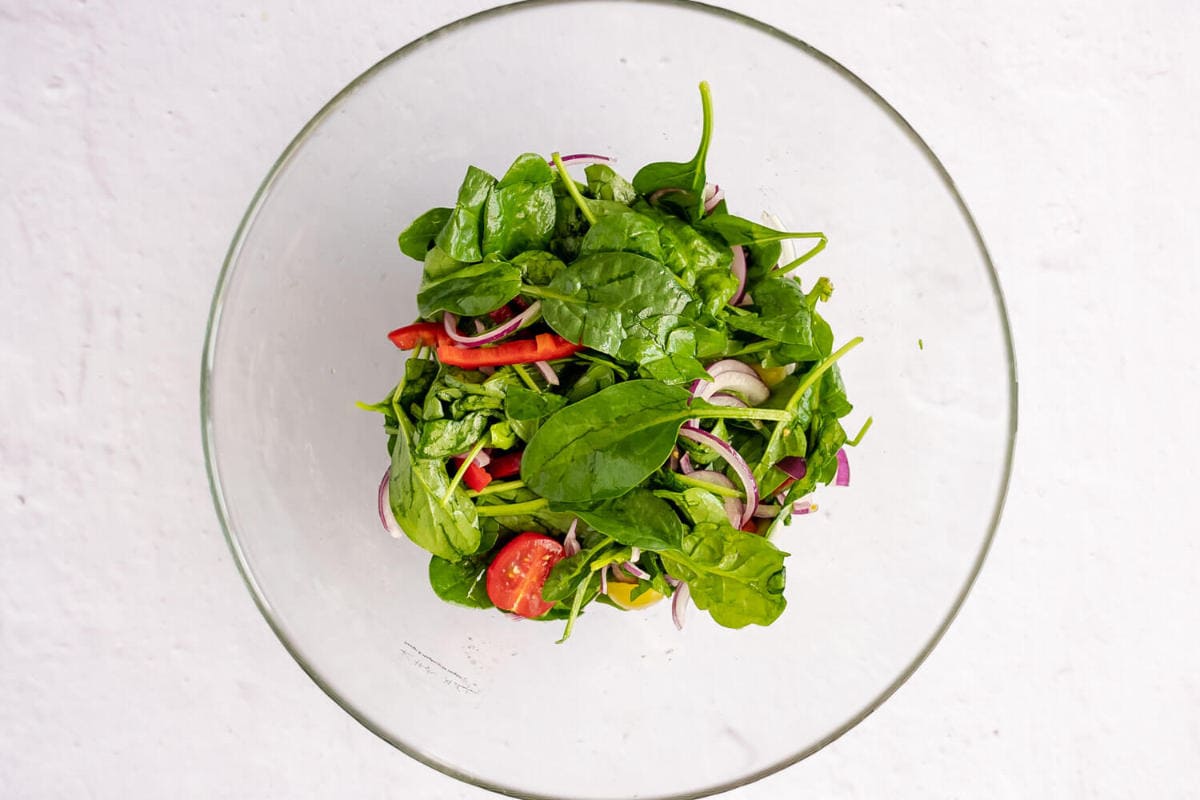 A glass bowl filled with fresh spinach leaves, sliced red onion, cherry tomatoes, and red bell pepper creates the perfect base for a vibrant grilled steak salad on a light, textured surface.