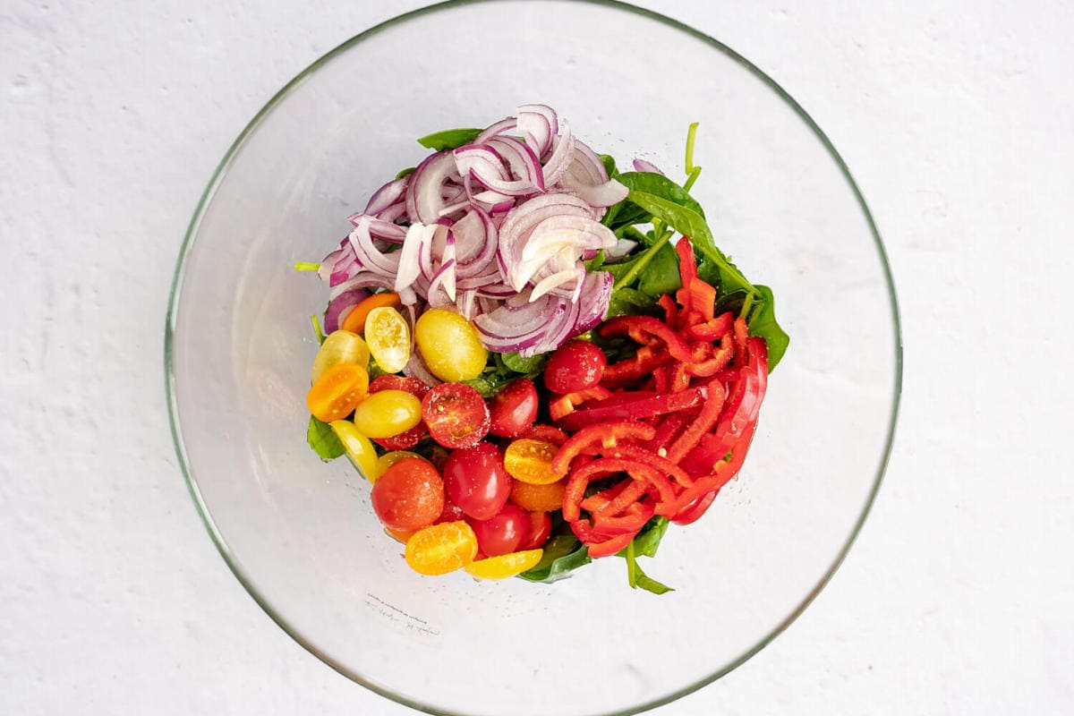 A glass bowl containing sliced red onions, halved cherry tomatoes, sliced red bell peppers, and spinach leaves arranged in separate sections, perfect as a fresh base for a vibrant grilled steak salad, on a white background.