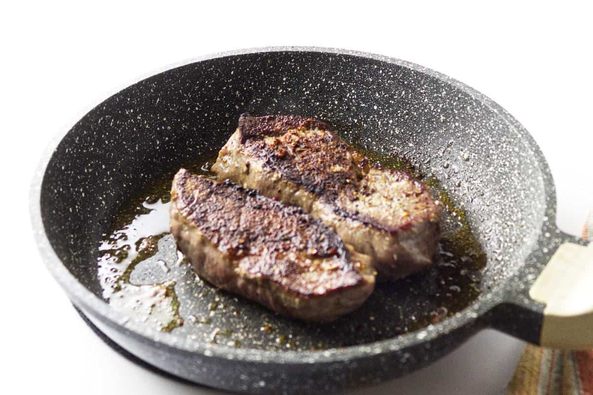 Two pieces of steak with a browned crust are cooking in a nonstick frying pan with oil, perfect for a grilled steak salad, set against a white background.