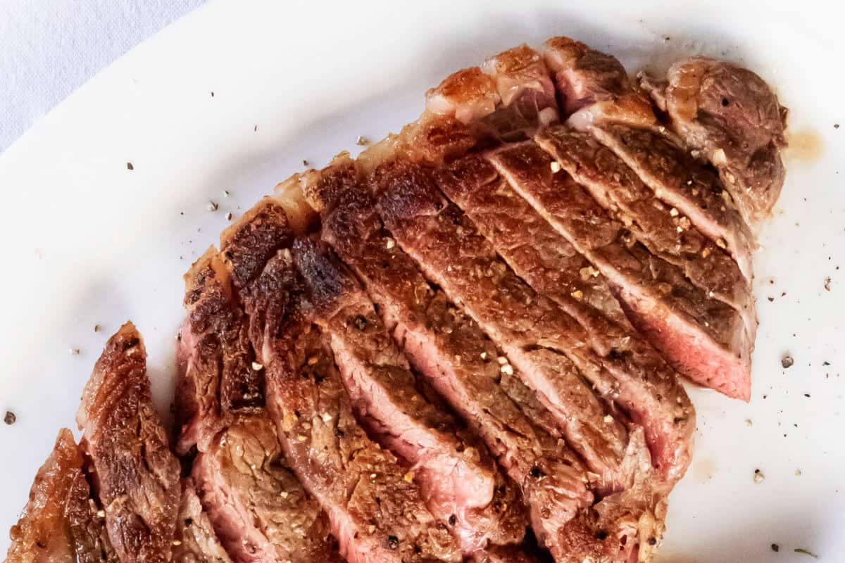 Sliced medium-rare grilled steak with a browned, seared crust, sprinkled with coarse salt and black pepper, served atop a fresh salad on a white plate.