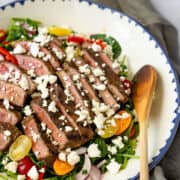 A grilled steak salad topped with slices of medium-rare steak and crumbled feta cheese, plus cherry tomatoes, radishes, and greens, served in a white bowl with a blue rim beside a wooden spoon.