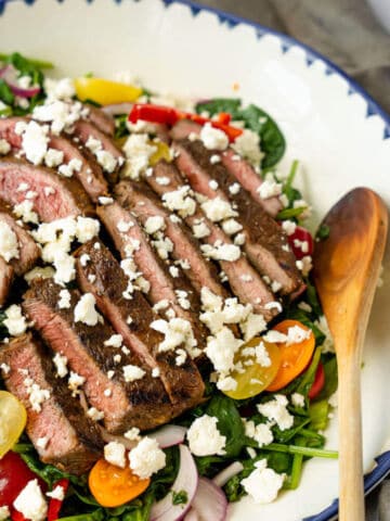 A grilled steak salad topped with slices of medium-rare steak and crumbled feta cheese, plus cherry tomatoes, radishes, and greens, served in a white bowl with a blue rim beside a wooden spoon.
