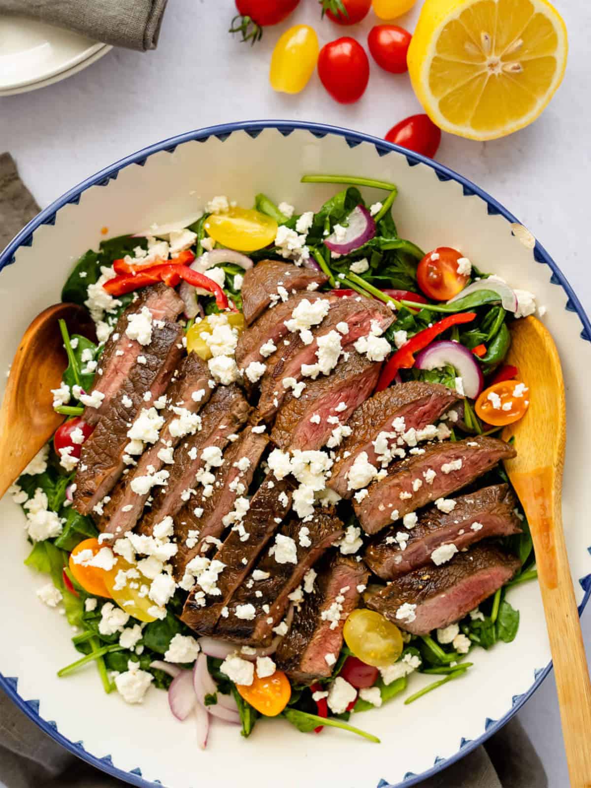 A grilled steak salad topped with crumbled feta cheese sits in a bowl, filled with spinach, cherry tomatoes, red onions, and red bell peppers. Lemon and tomatoes rest beside the bowl, while wooden salad utensils are inside.