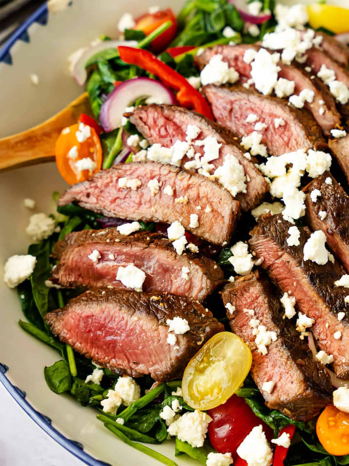 A close-up of a grilled steak salad topped with crumbled feta, cherry tomatoes, spinach, red onions, and bell peppers, served in a bowl with a wooden spoon.