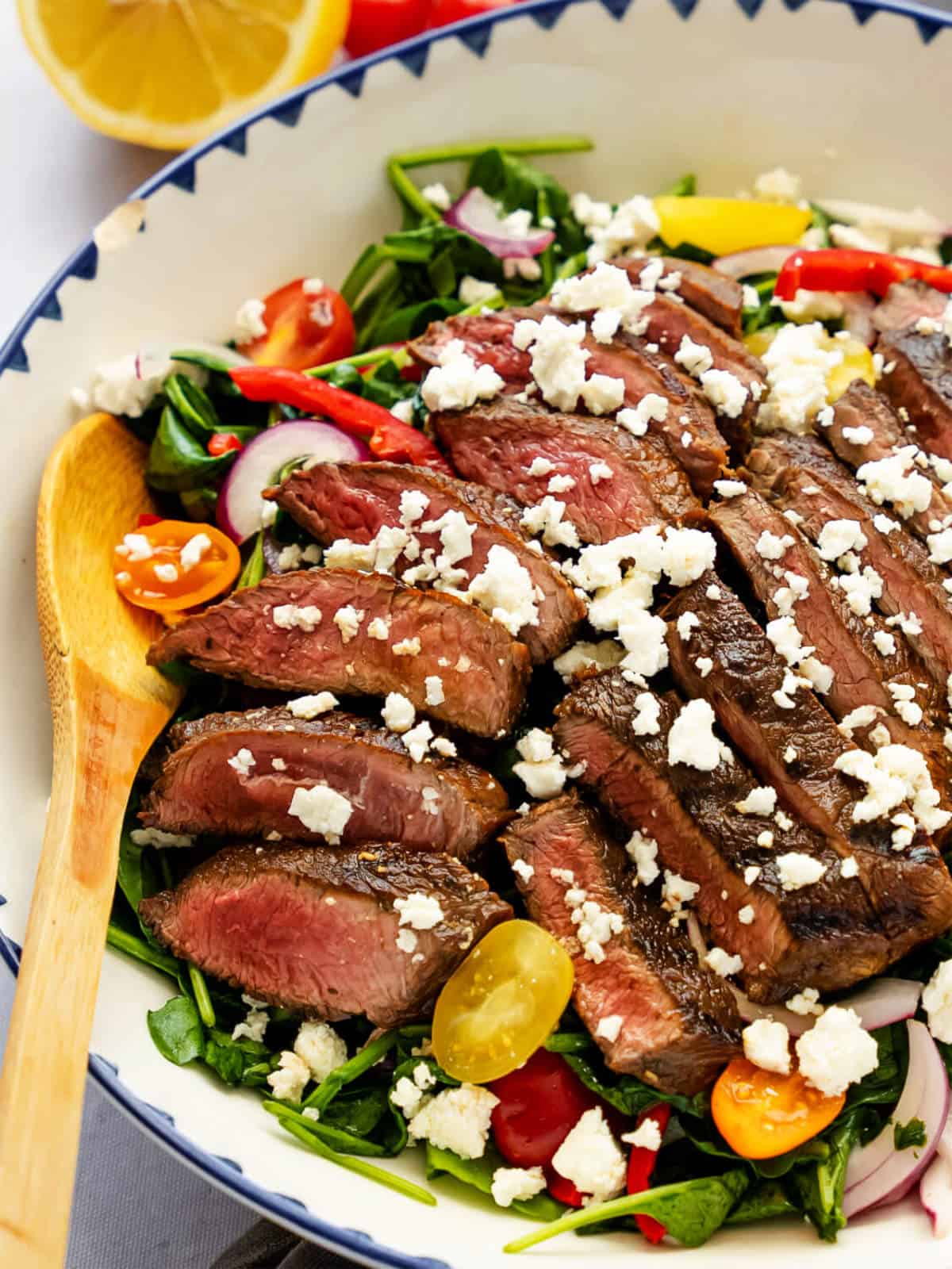 A grilled steak salad bowl filled with sliced medium-rare steak, cherry tomatoes, spinach, red onions, and crumbled feta cheese, with a wooden serving spoon on the side.