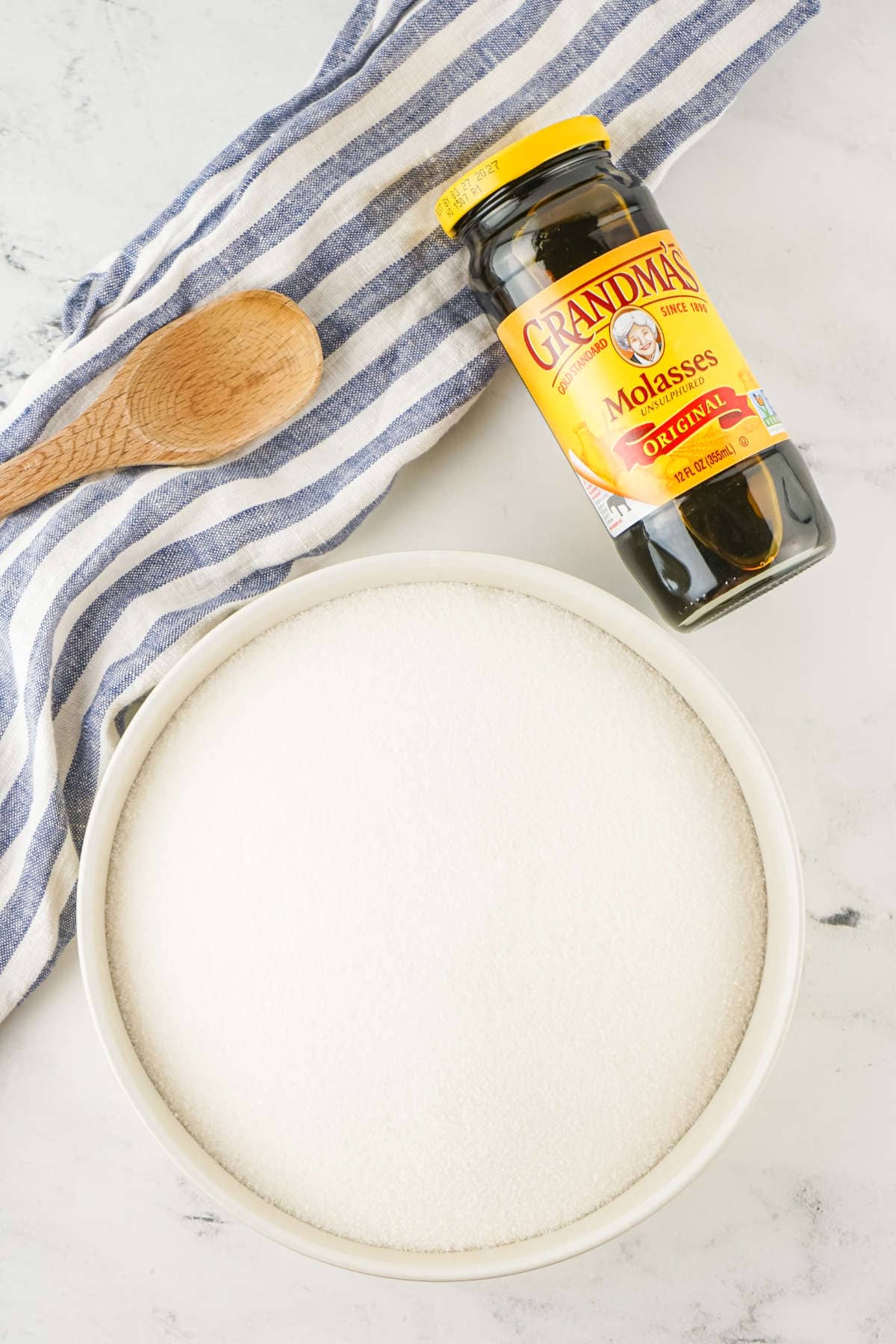 A bowl of homemade brown sugar substitute, a jar of Grandma’s Original Molasses, a wooden spoon, and a blue-and-white striped cloth rest on a marble countertop.
