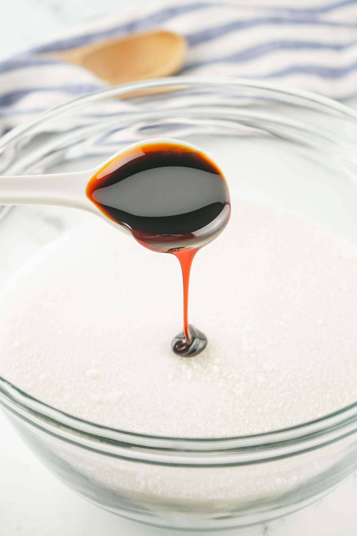 A spoonful of dark liquid, likely molasses, is being poured over a bowl filled with granulated white sugar-a classic way to create a homemade brown sugar substitute. A striped cloth and a wooden spoon are blurred in the background.