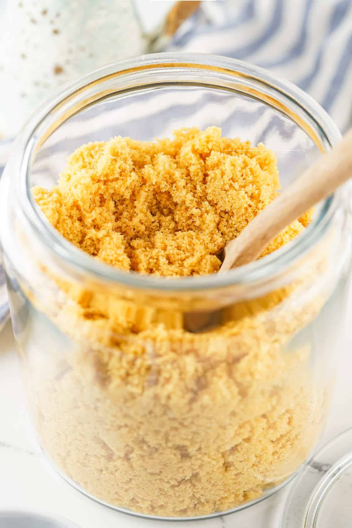 A clear glass jar filled with homemade brown sugar substitute, with a wooden spoon inside. The jar is open and resting on a white surface with a blue and white striped cloth in the background.