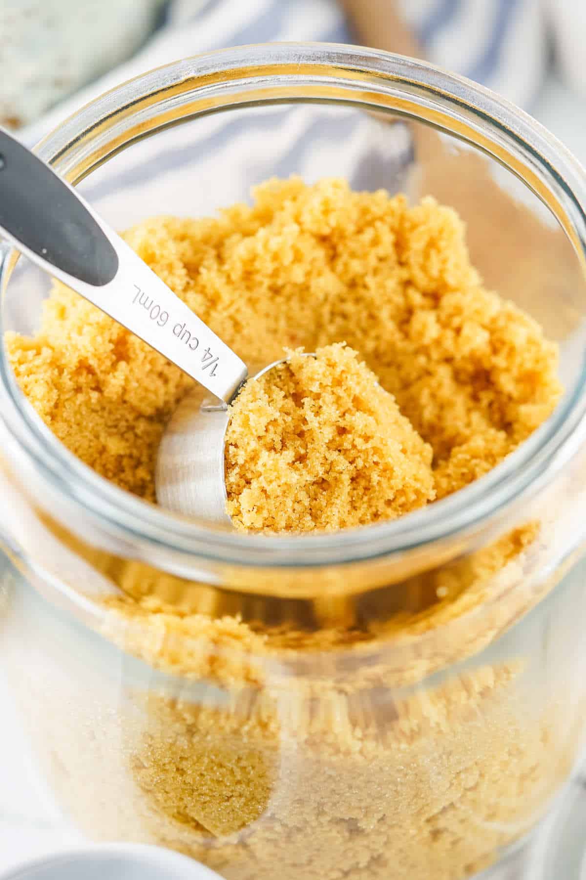 A close-up of a glass jar filled with homemade brown sugar substitute, with a metal ¼ cup measuring scoop partially submerged in the sugar.