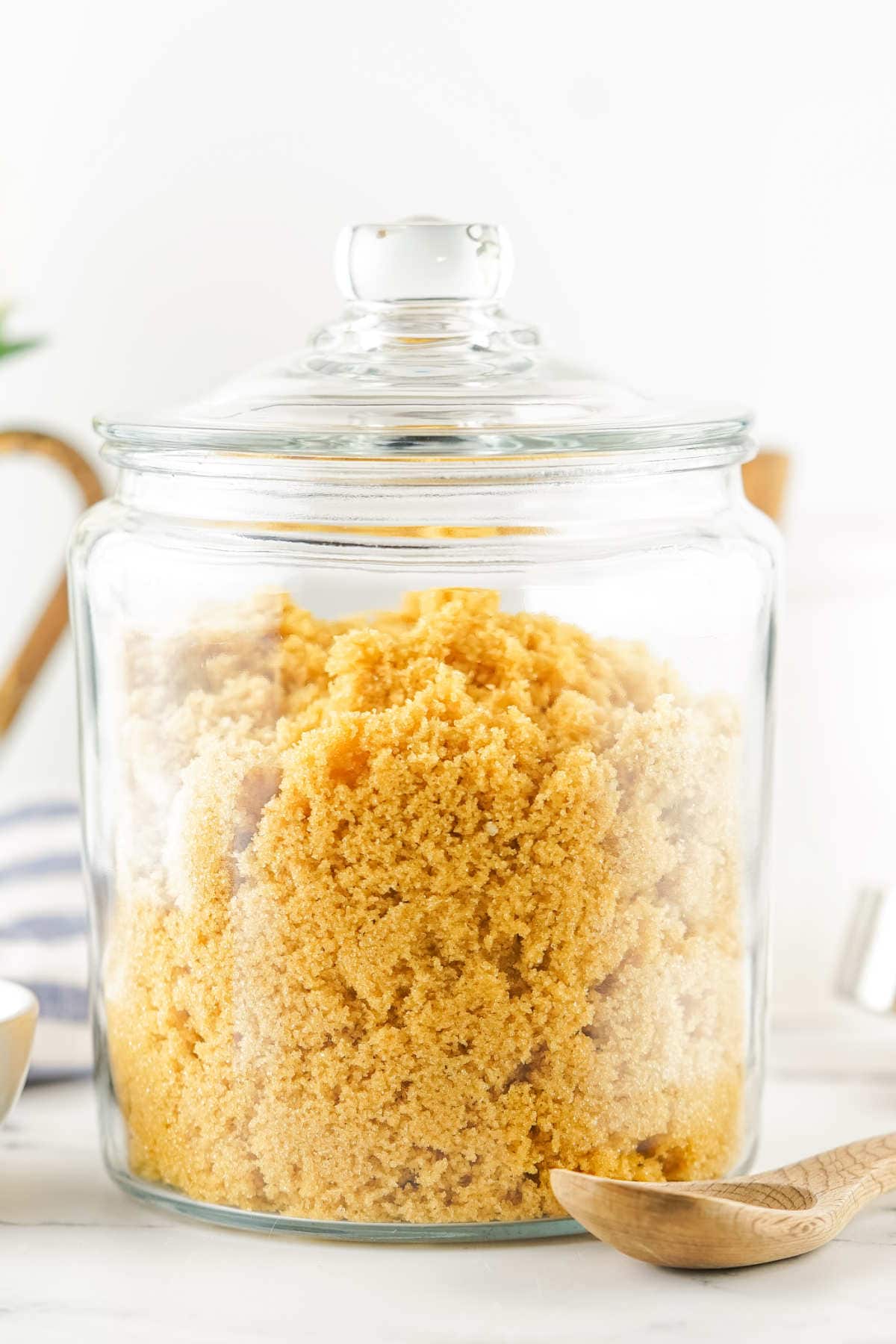 A clear glass jar filled with homemade brown sugar substitute sits on a white surface. The jar has a matching glass lid, and a wooden spoon rests nearby. The background is bright and softly focused.