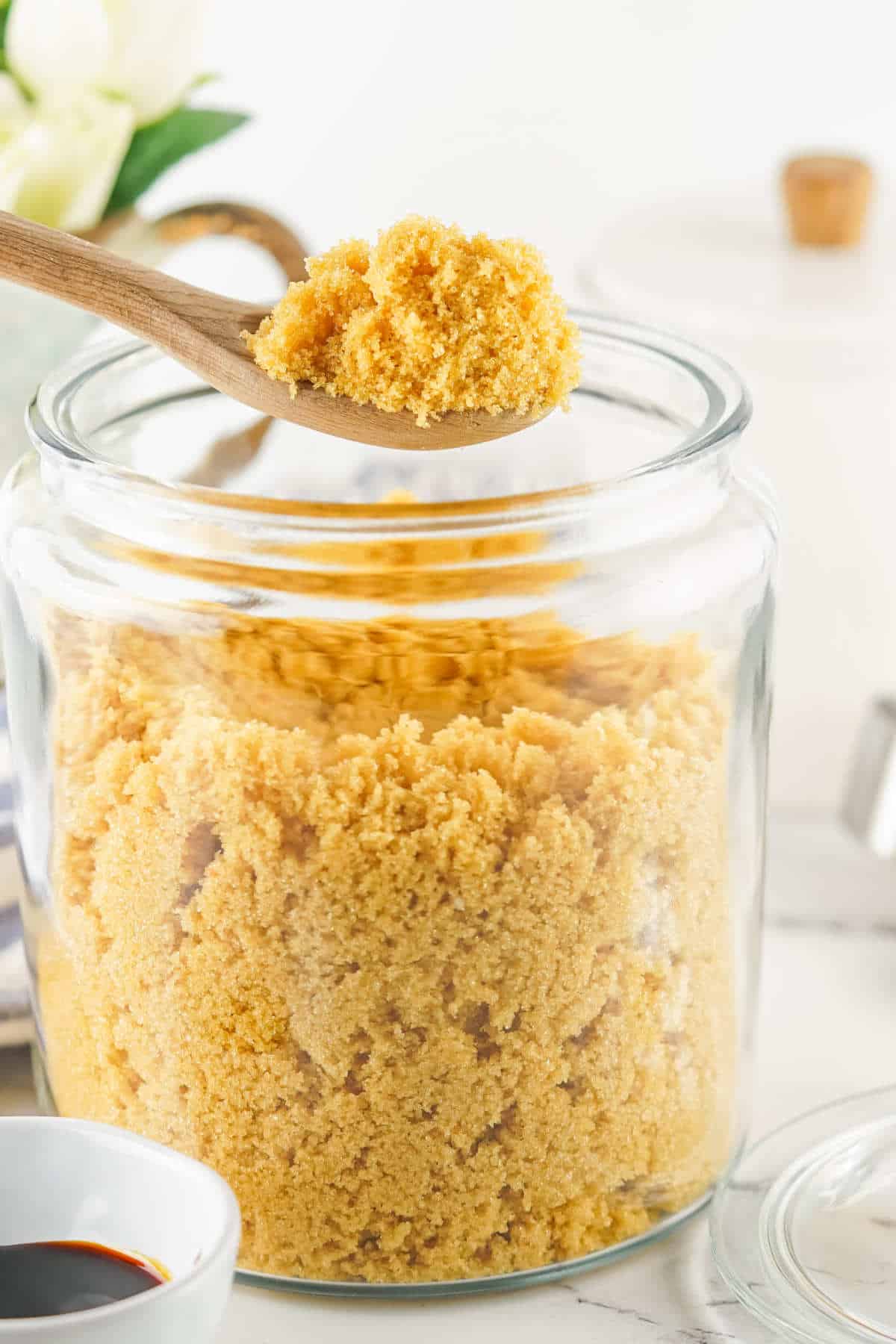 A glass jar filled with homemade brown sugar substitute sits on a countertop. A wooden spoon holds a scoop of the sugar above the jar. In the foreground, a small bowl with dark liquid is partially visible.