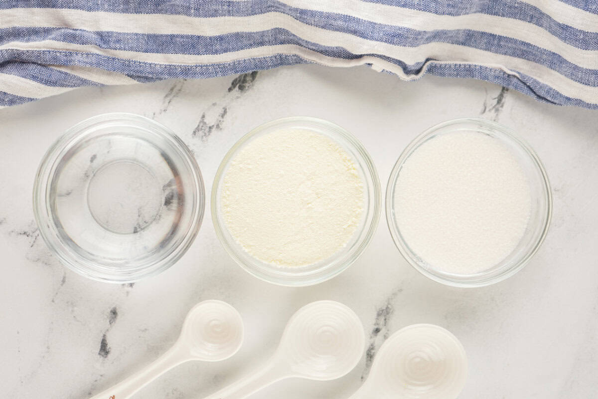 Three glass bowls containing water, powdered milk, and sugar—ingredients for homemade sweetened condensed milk—are arranged in a row on a marble surface next to a blue and white striped cloth. White measuring spoons are placed below the bowls.