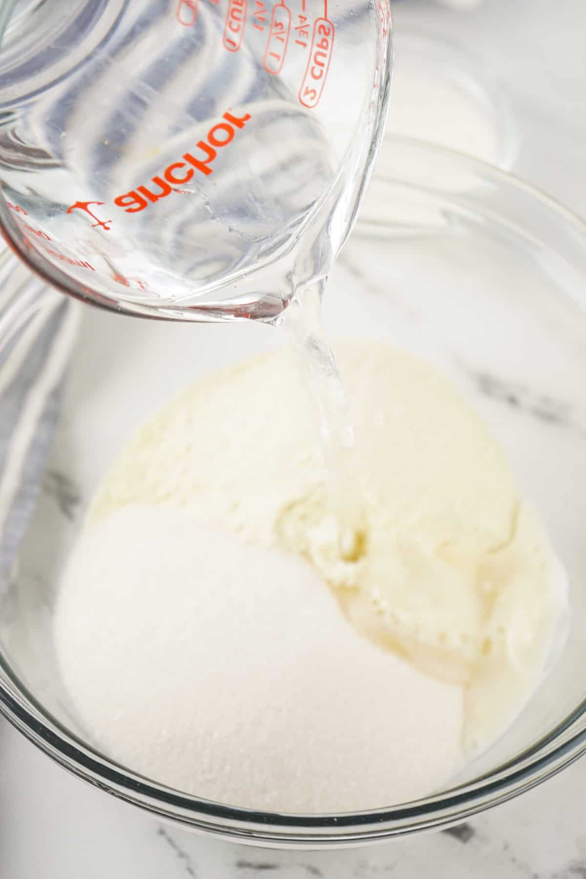 A glass measuring cup pours water into a clear mixing bowl containing sugar and a creamy homemade sweetened condensed milk mixture, all set on a white marble surface.