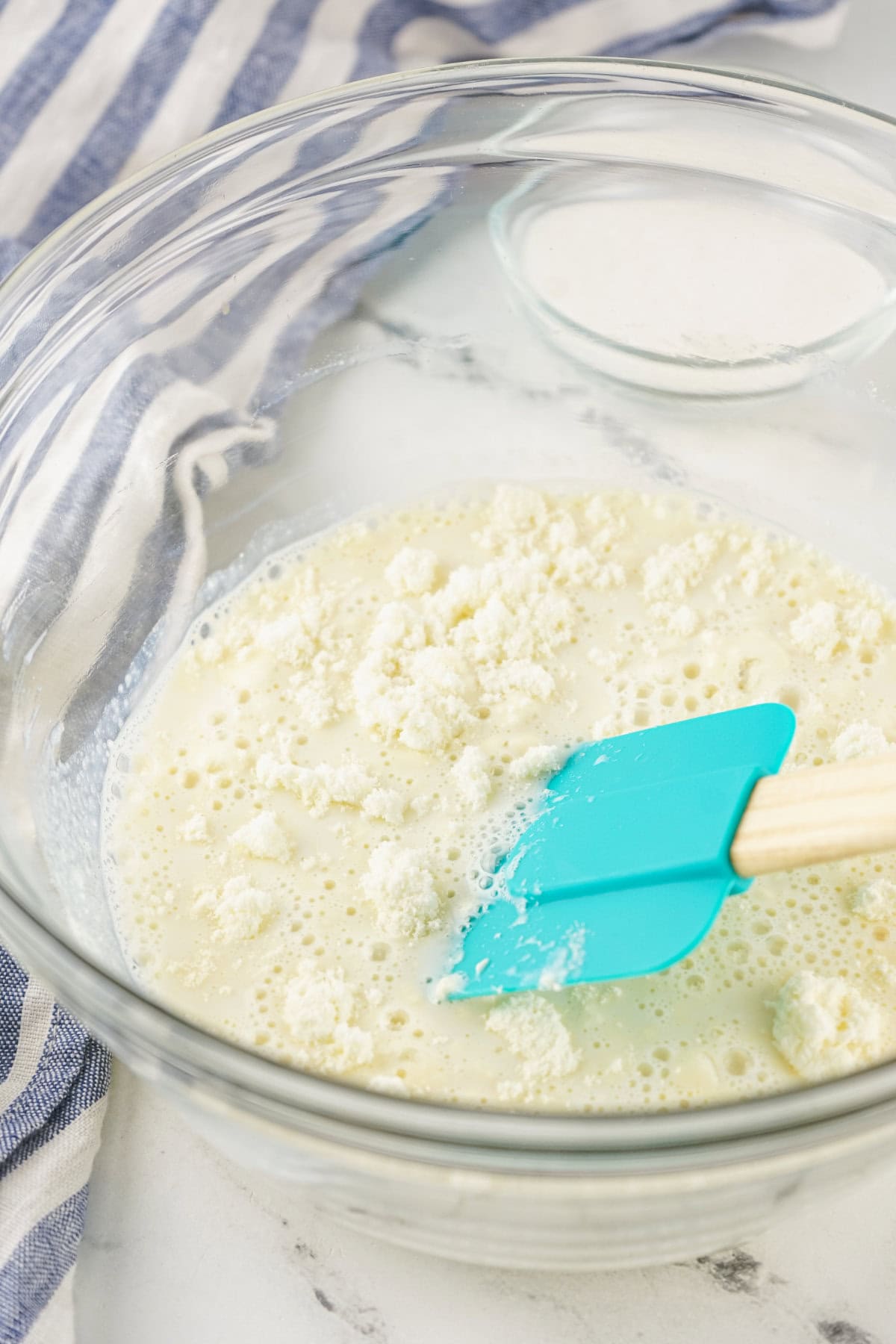 A glass mixing bowl with milk, crumbled cheese, and homemade sweetened condensed milk, stirred with a blue spatula, sits on a marble surface next to a small bowl of sugar and a striped cloth.
