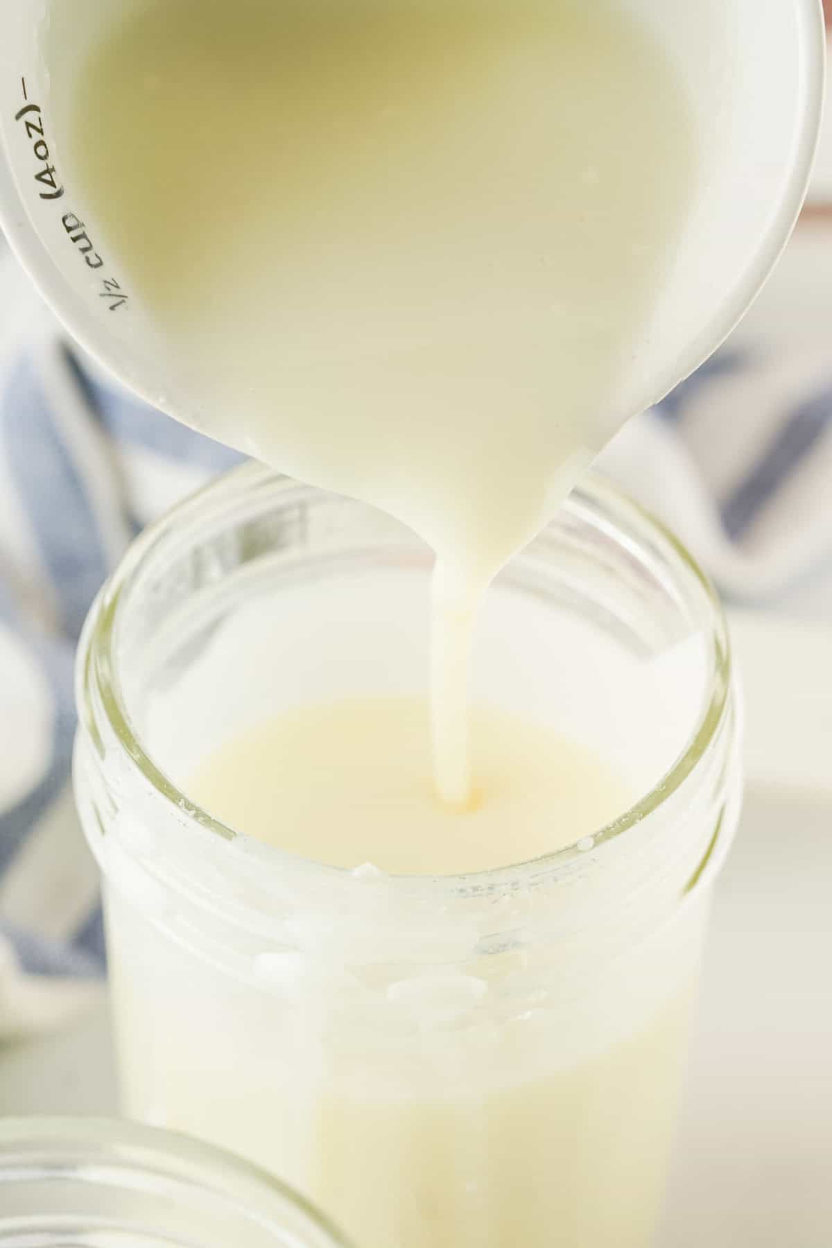 A close-up of homemade sweetened condensed milk being poured from a measuring cup into a glass jar, with a blurred striped cloth in the background.