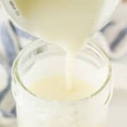 A close-up of thick, creamy homemade sweetened condensed milk being poured from a measuring cup into a glass jar, with a blue and white striped cloth in the background.