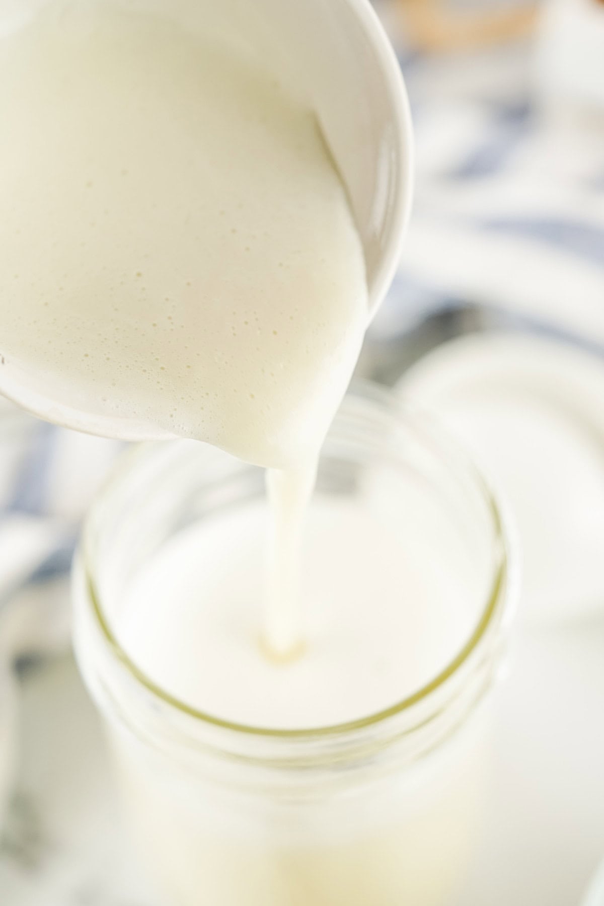 A creamy white liquid of homemade sweetened condensed milk is being poured from a small bowl into a glass jar, with a soft-focus background.