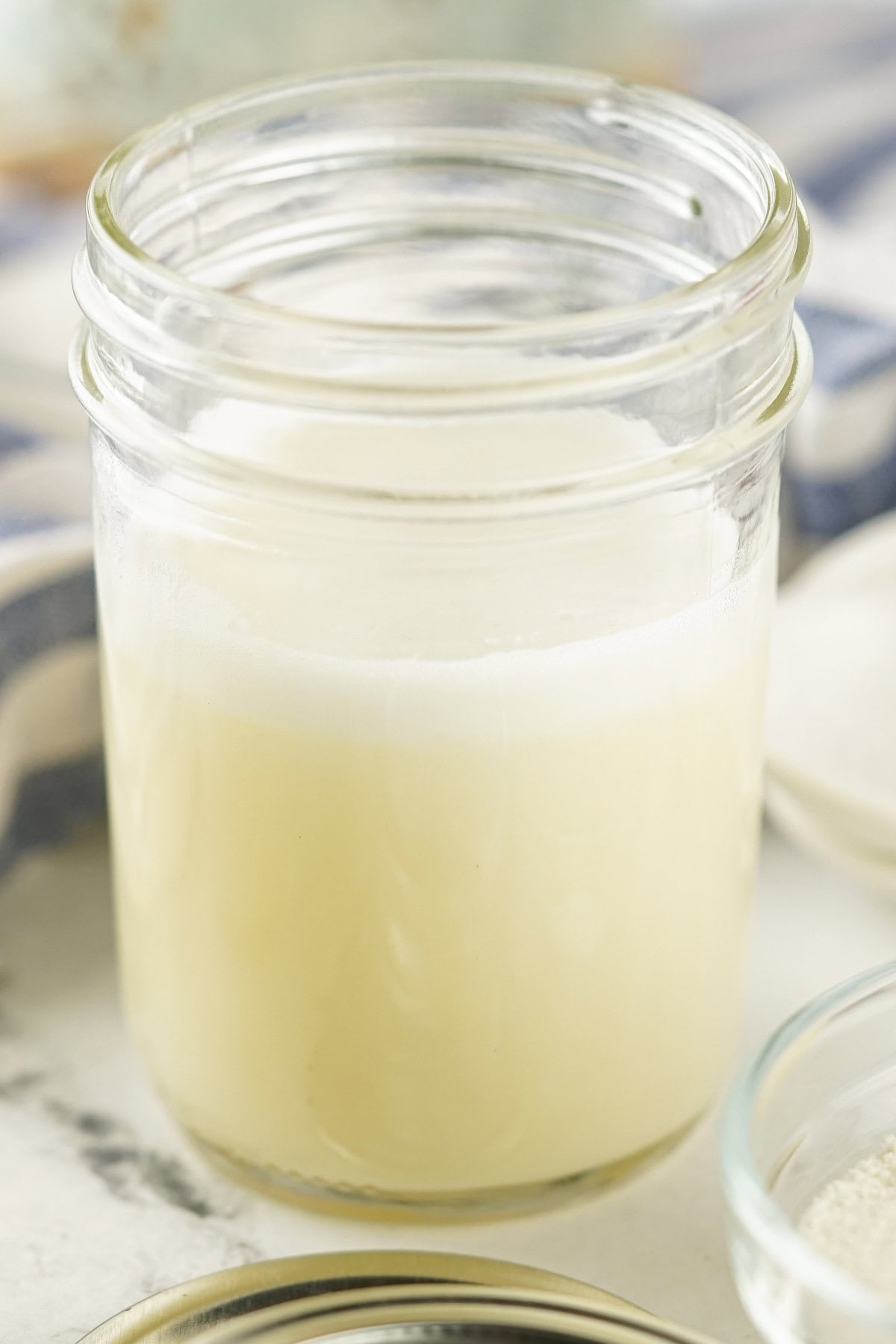 A clear glass jar filled with creamy, homemade sweetened condensed milk sits on a white surface, with a blue and white striped cloth in the background.